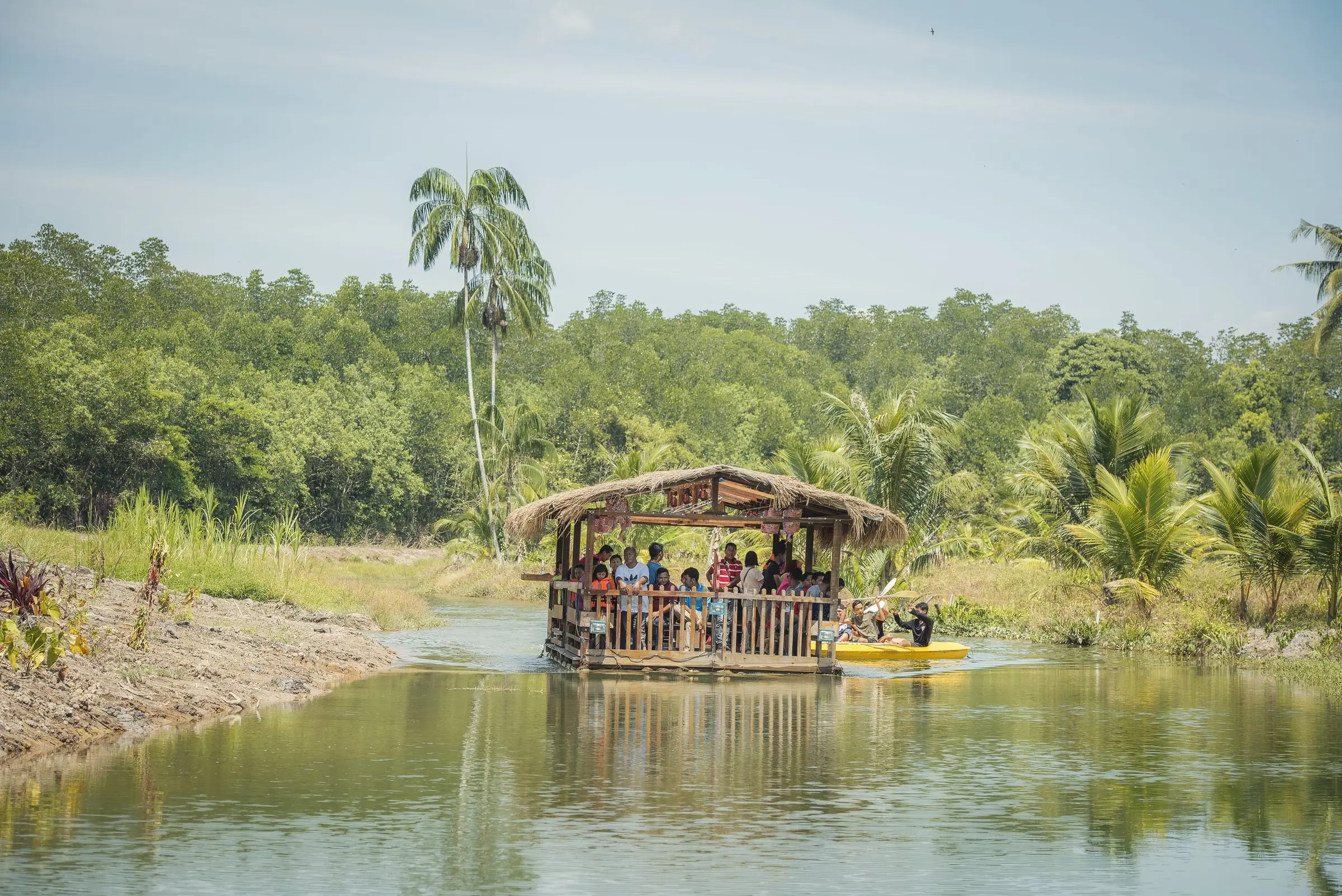 Guests aboard a thatched-roof River Cruise boat on a calm, tree-lined river