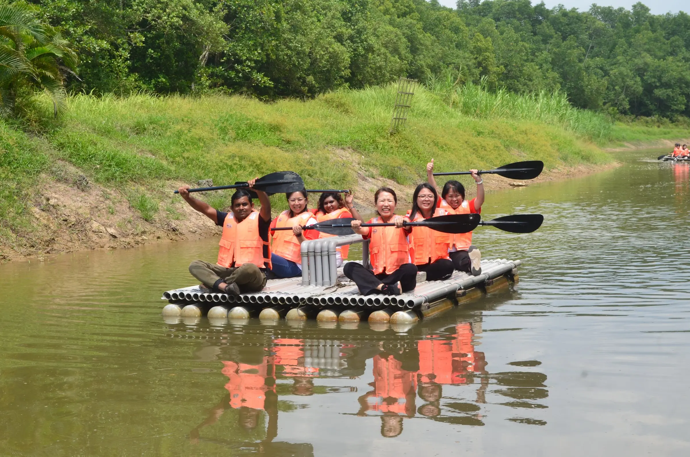 A group in orange life jackets cheers with paddles raised on a pipe raft on the river