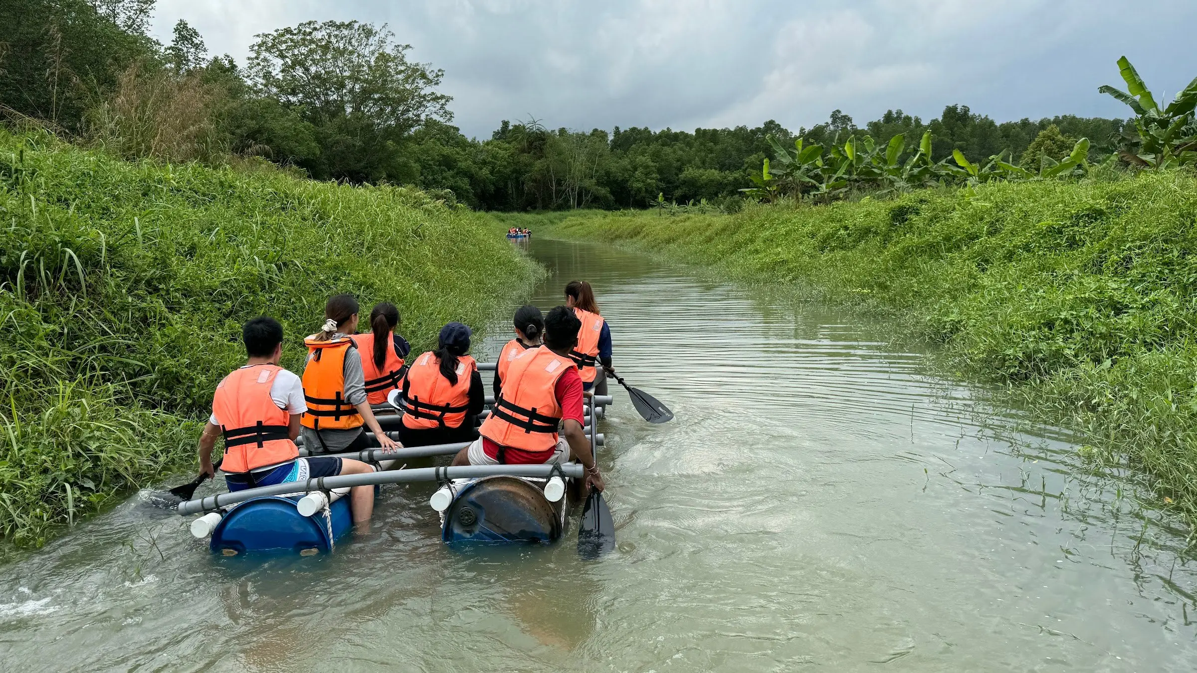 Two teams paddle home-built barrel rafts down a narrow green river, seen from behind