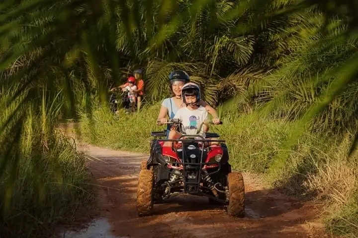An adult and child ride an ATV out of a shaded oil palm trail
