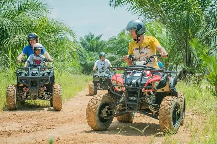 Riders in helmets drive muddy ATVs along a dirt trail through the oil palms