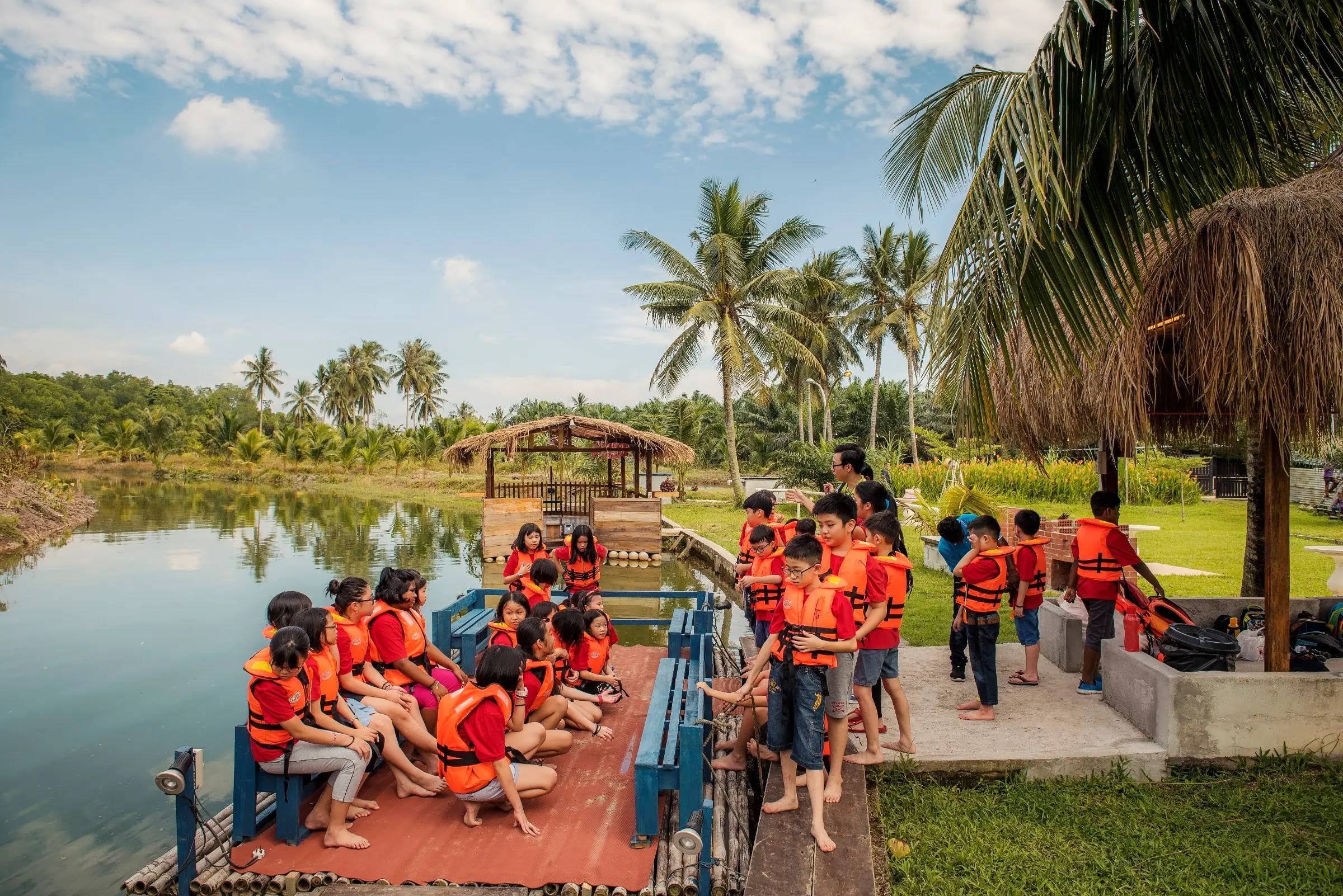 Children in orange life jackets wait on a lakeside jetty beside a thatched gazebo and palms