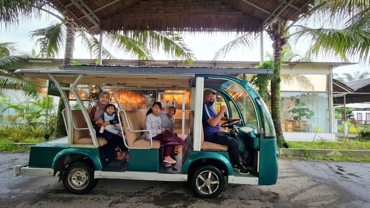 A family rides the resort’s green shuttle buggy car, with a driver, past the thatched hall