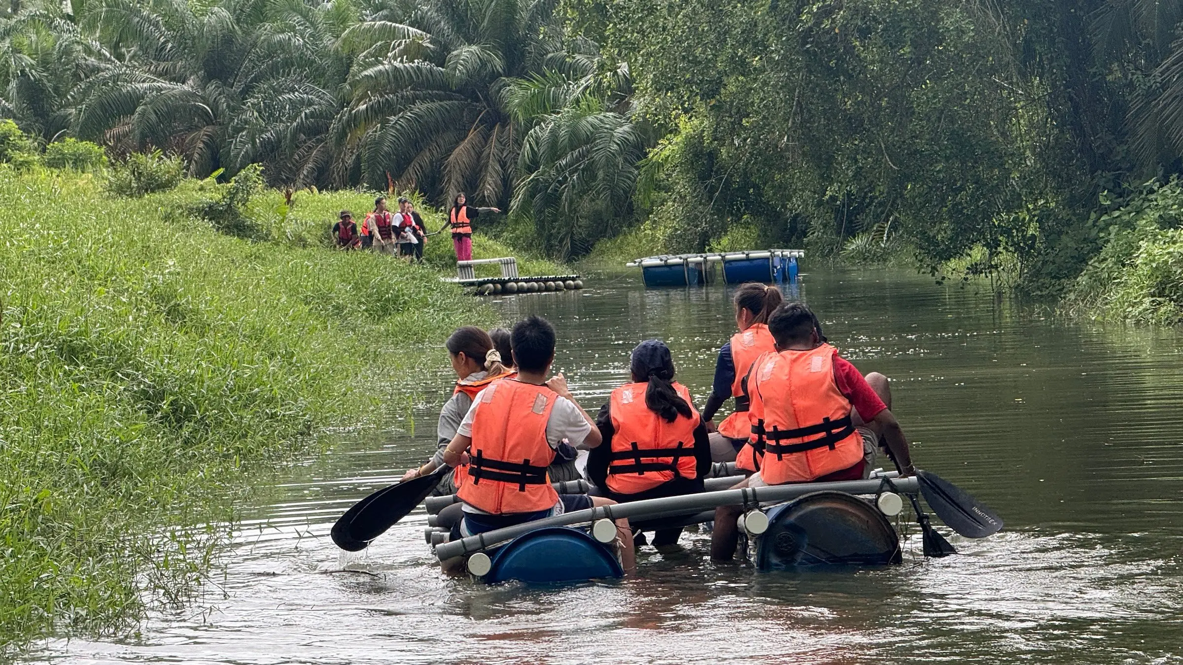 Colleagues in life jackets paddle a barrel raft down a palm-lined river, seen from behind