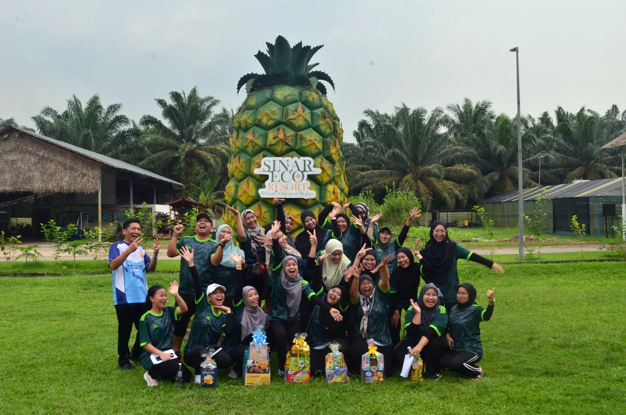 A big group cheers together on the lawn in front of the giant pineapple Sinar Eco sign
