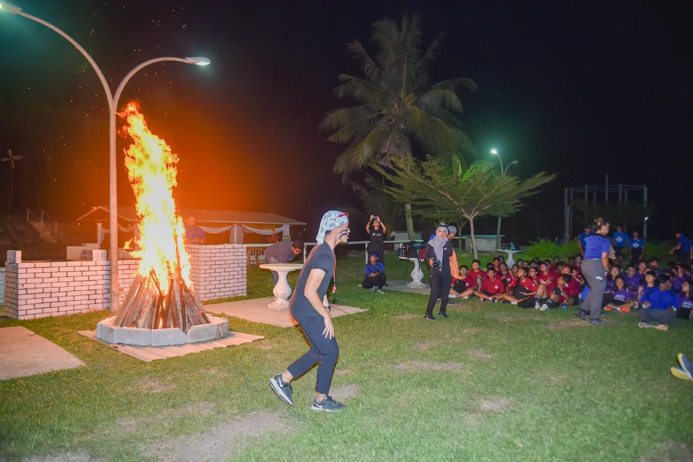 A bonfire blazes on the lawn at night while a large group sits together nearby