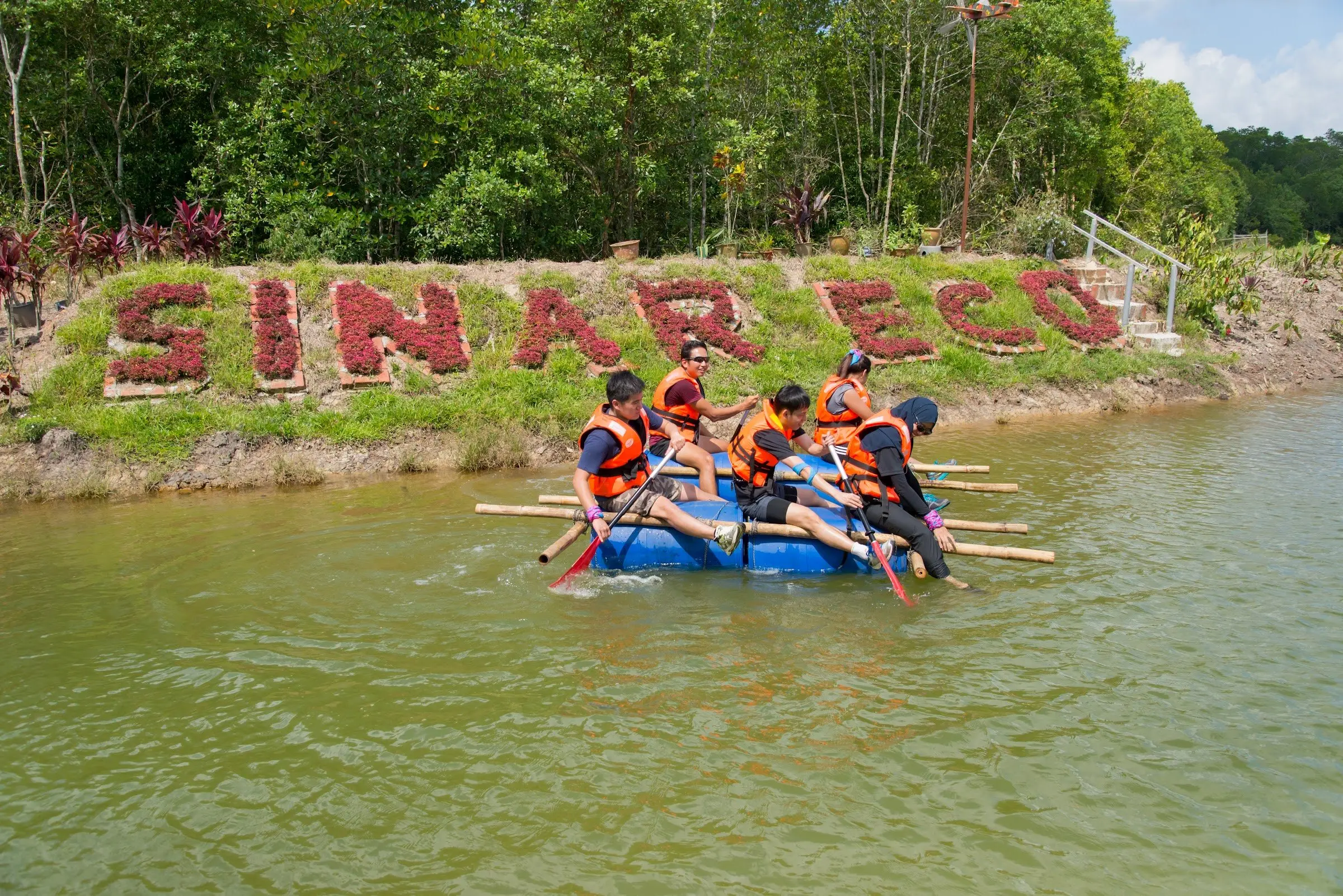 Students paddle a raft past the flower-planted SINAR ECO sign on the riverbank