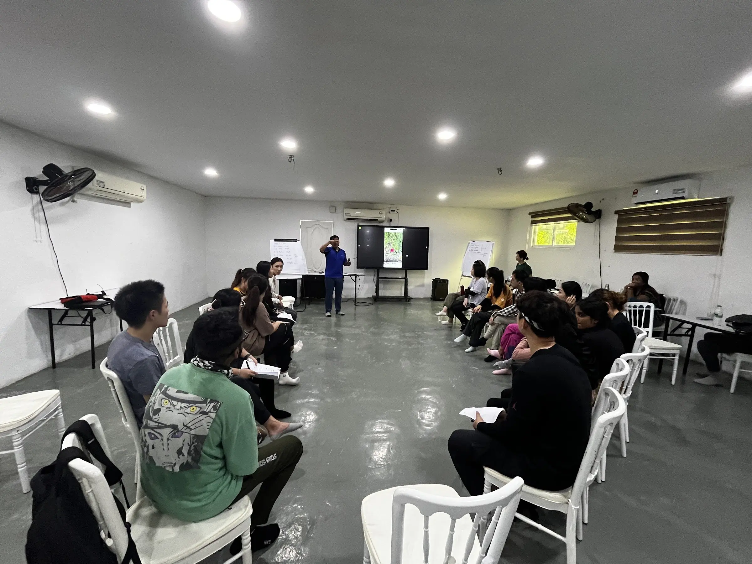 A team sits in a U-shape in the meeting hall while a presenter speaks beside the screen