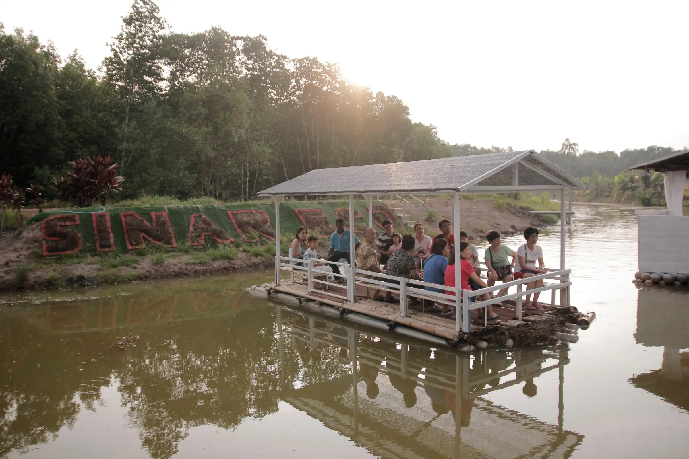 A covered River Cruise raft full of guests drifts past the SINAR ECO sign at golden hour