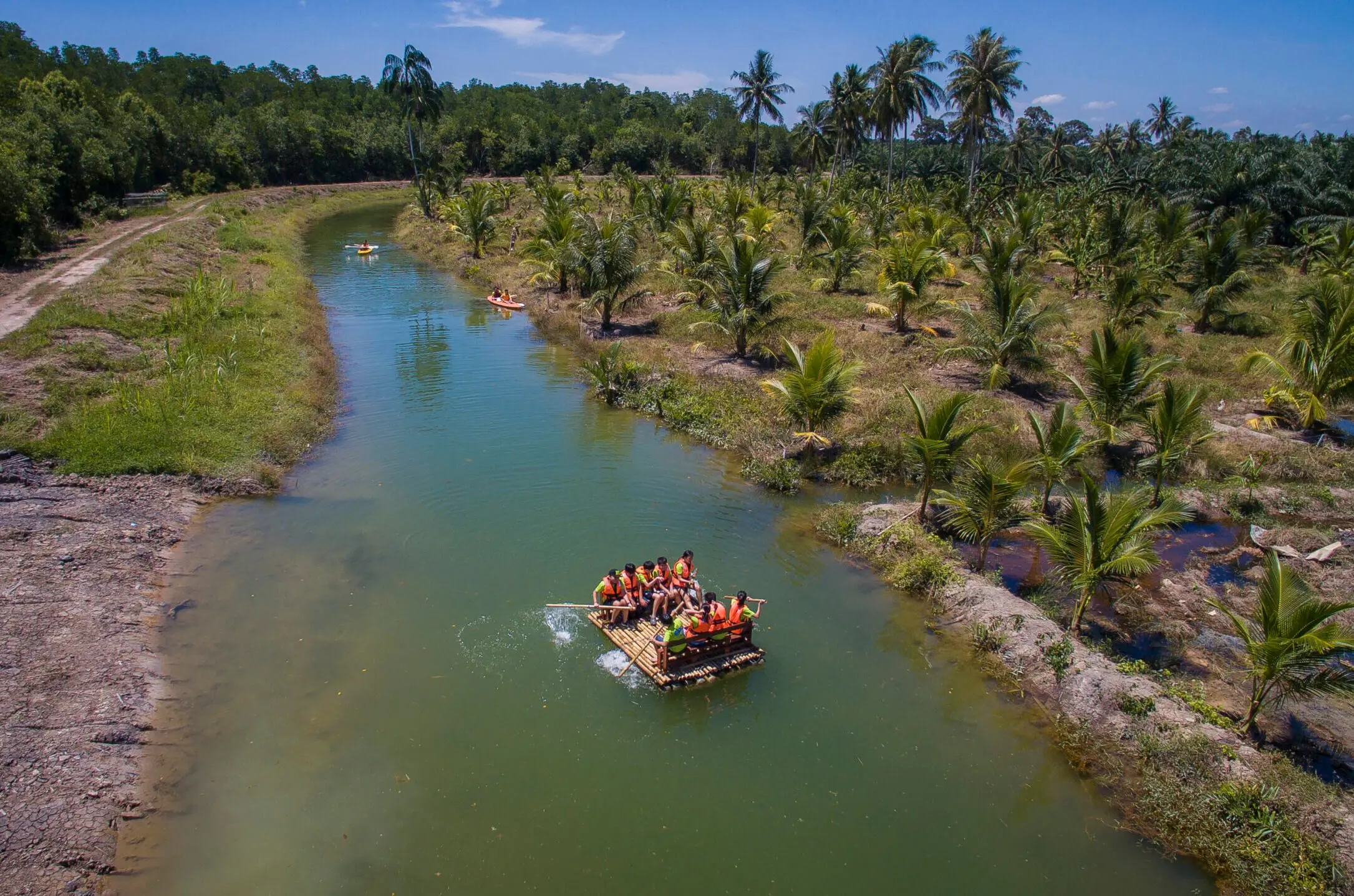 Aerial view of a raft drifting down a calm green river between rows of young palms